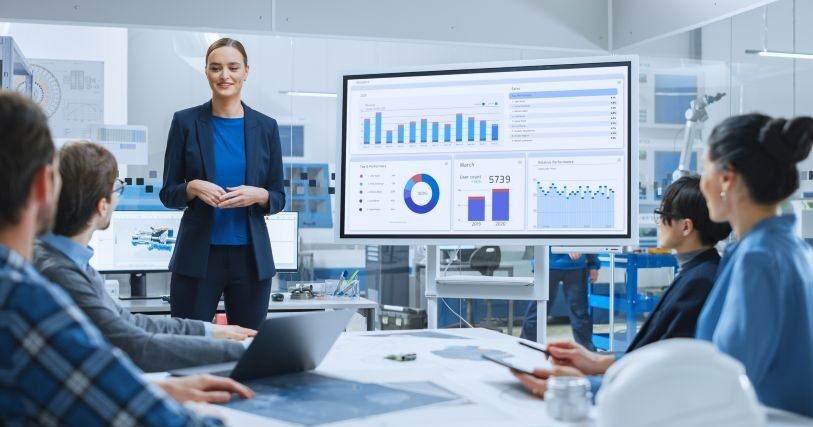 A young woman in a dark blue suit stands next to a large chart and explains to 4 colleagues what the data show.