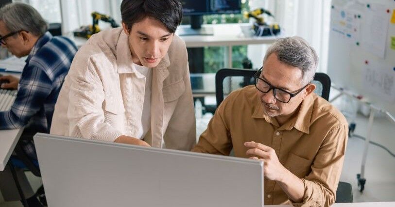 Two Asian men cluster near a computer screen; one watches as the other points to something he is explaining.