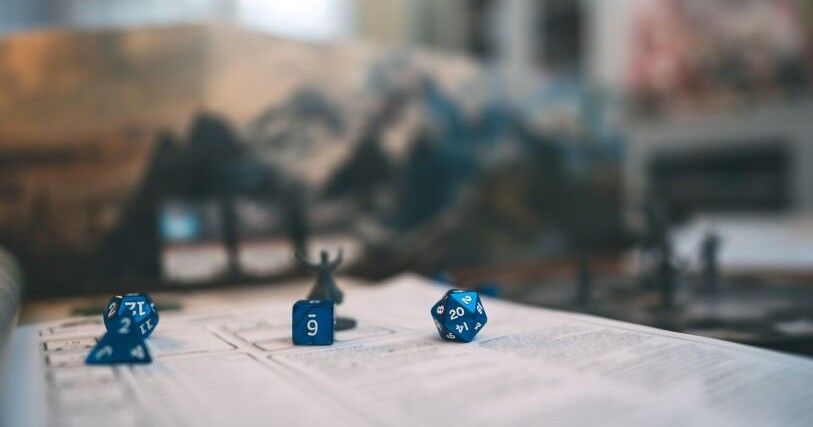 Blue game dice rest on a piece of paper with game instructions with blurry gamepieces in the background
