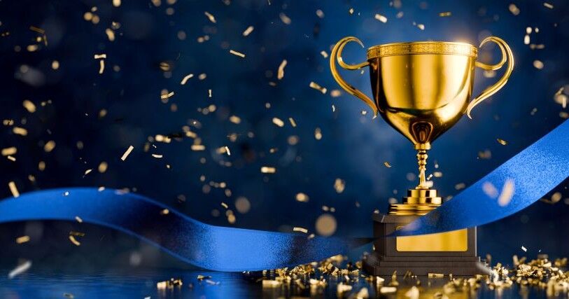 A large gold trophy cup, with a blue ribbon floating around it signifies winning. It stands against a dark background with gold confetti falling around it.