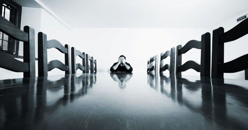 A person sits at the head of a long table, with empty chairs along both sides. Their head in in their hands, signaling frustration or overwhelm. The image in in shades of gray.